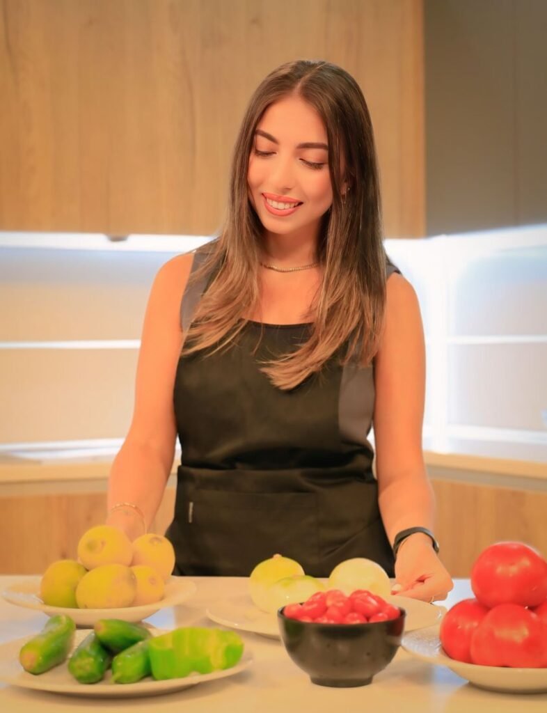 Nour, founder of Nourrition, smiling over a counter of fresh fruit and vegetables